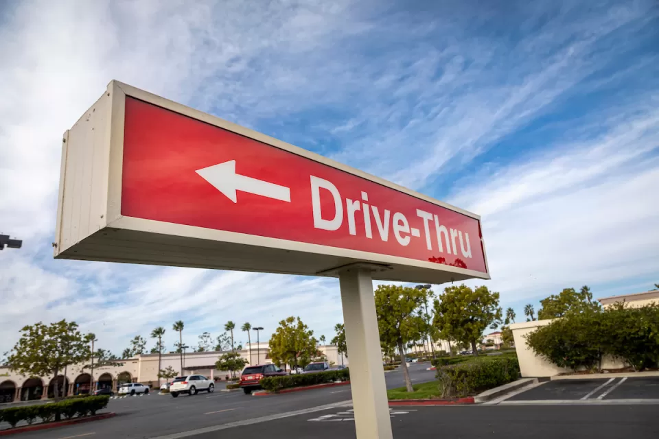 Close-up of red sign with white "Drive-Thru" text and arrow pointing left against partly cloudy blue sky.